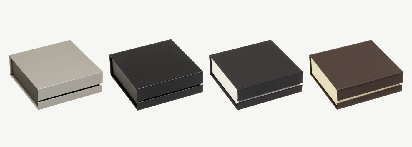 Vier geschlossene flache Klappschachteln nebeneinander, 80 × 80 × 25 mm, in Silber/Schwarz, Schwarz/Schwarz, Schwarz/Weiß und Braun/Creme