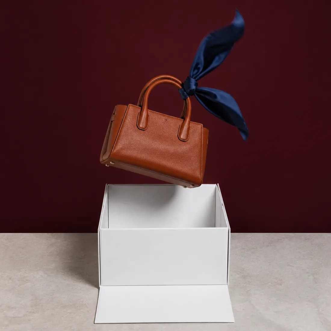 Braune Ledertasche mit dunkelblauem Seidentuch über einer geöffneten weißen Magnetbox vor bordeauxrotem Hintergrund