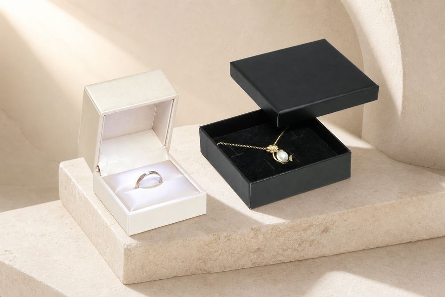 Links ein cremefarbenes Klappetui mit Ring, rechts eine flache schwarze Schmuckschachtel mit Kette